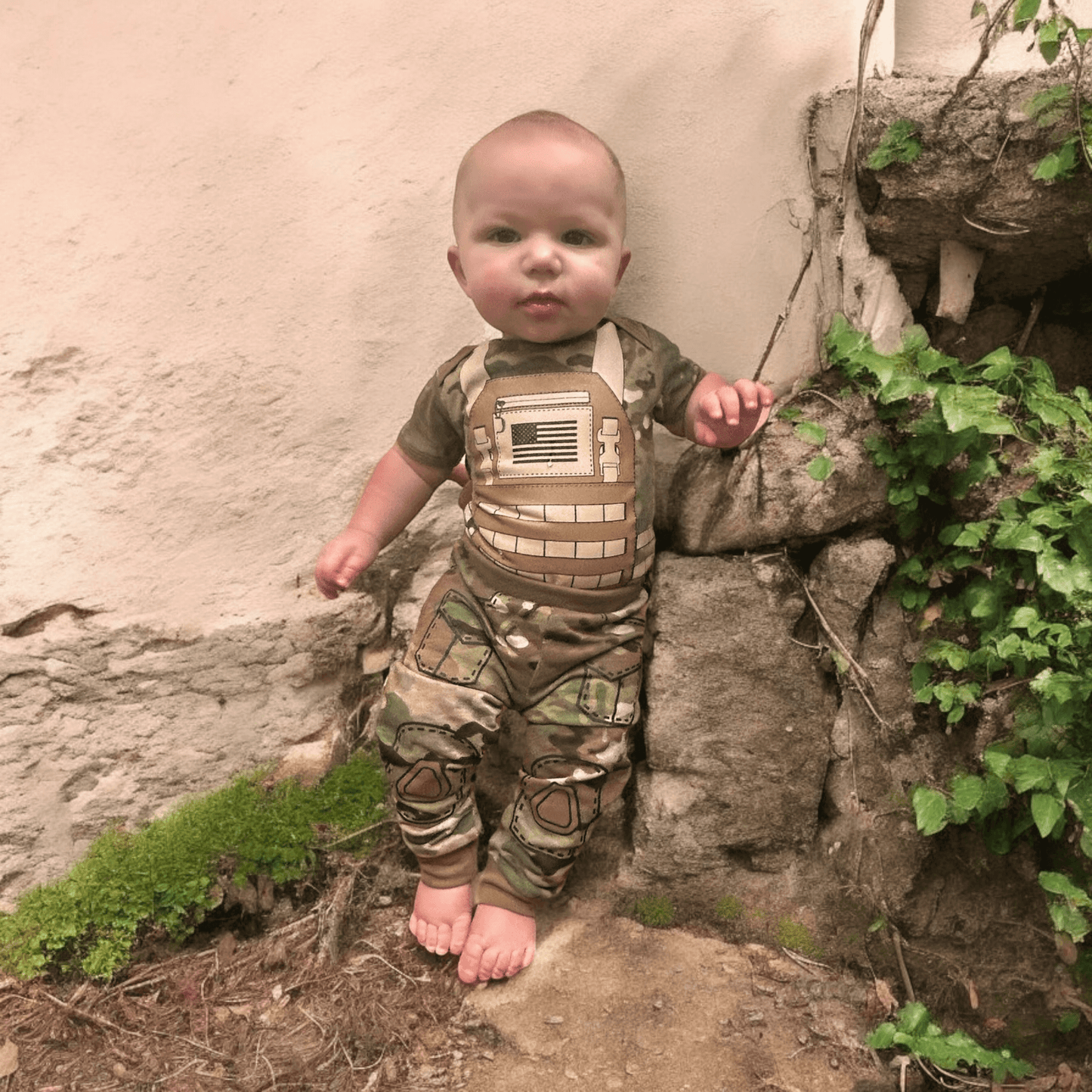 Baby wearing Multicam Tactical Combat Uniform with flak jacket design and US flag, standing outdoors.