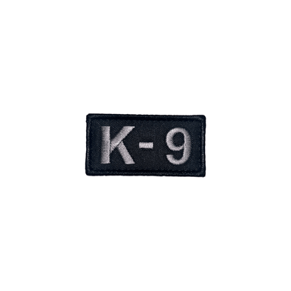 Embroidered patch featuring 'K-9' in light gray, perfect for dog vests, dimensions 2 3/4" x 1 3/8", with hook and loop.