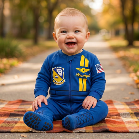 Baby wearing Blue Angels flight suit with patches, smiling on plaid blanket outdoors.